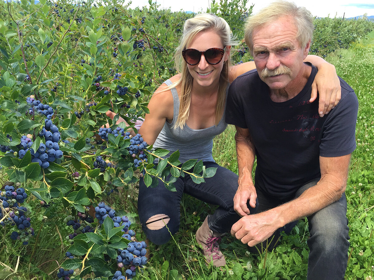 Two individuals, a woman and a man, are smiling while kneeling among lush blueberry bushes. The woman, wearing sunglasses and a tank top, holds a branch laden with ripe blueberries, while the man, dressed in a black shirt, poses beside her. The scene captures the vibrant greenery of the blueberry farm, highlighting the abundance of fruit and the joy of berry picking.