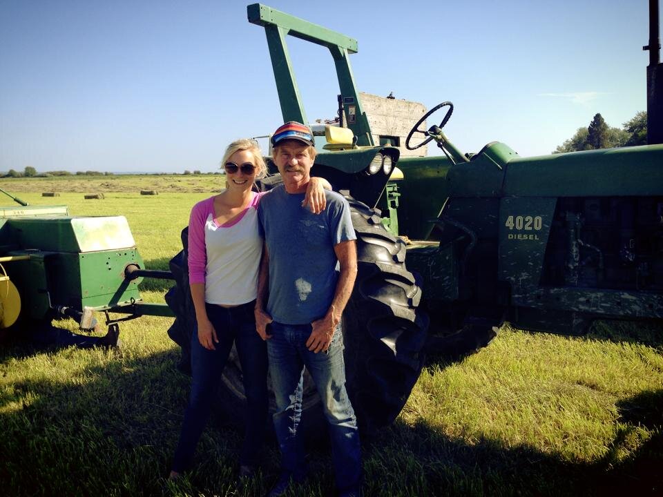 A young woman and an older man stand together beside a green John Deere tractor in a sunny field, showcasing a connection to farming and agriculture. The background features a clear blue sky and open grassland, emphasizing a rural lifestyle.