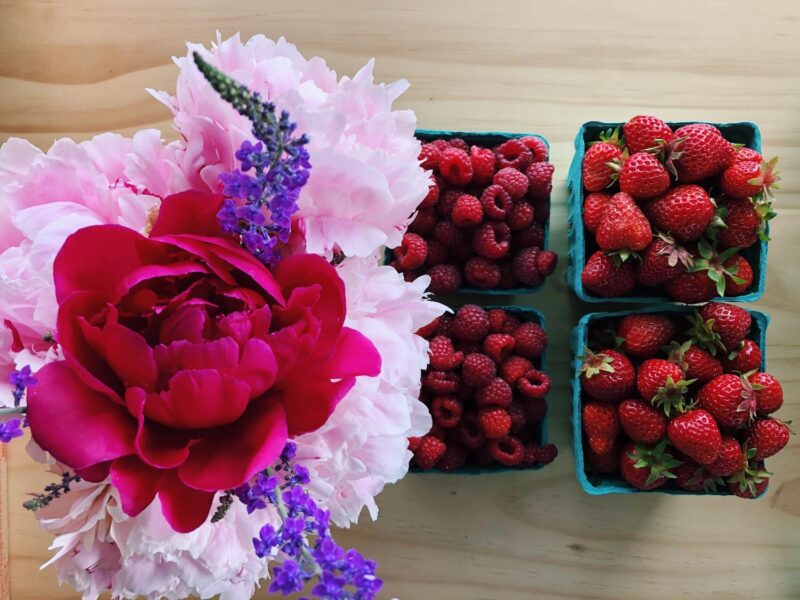 Fresh strawberries and raspberries in blue containers accompanied by a vibrant peony and lavender flowers on a wooden surface, showcasing a colorful and healthy arrangement.