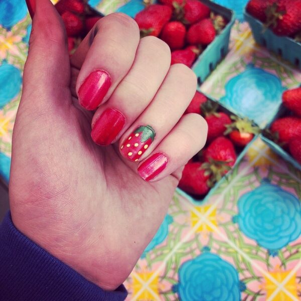 Close-up of a hand with vibrant red nail polish, featuring a strawberry design on one nail, set against a colorful background of fresh strawberries and a patterned tablecloth.