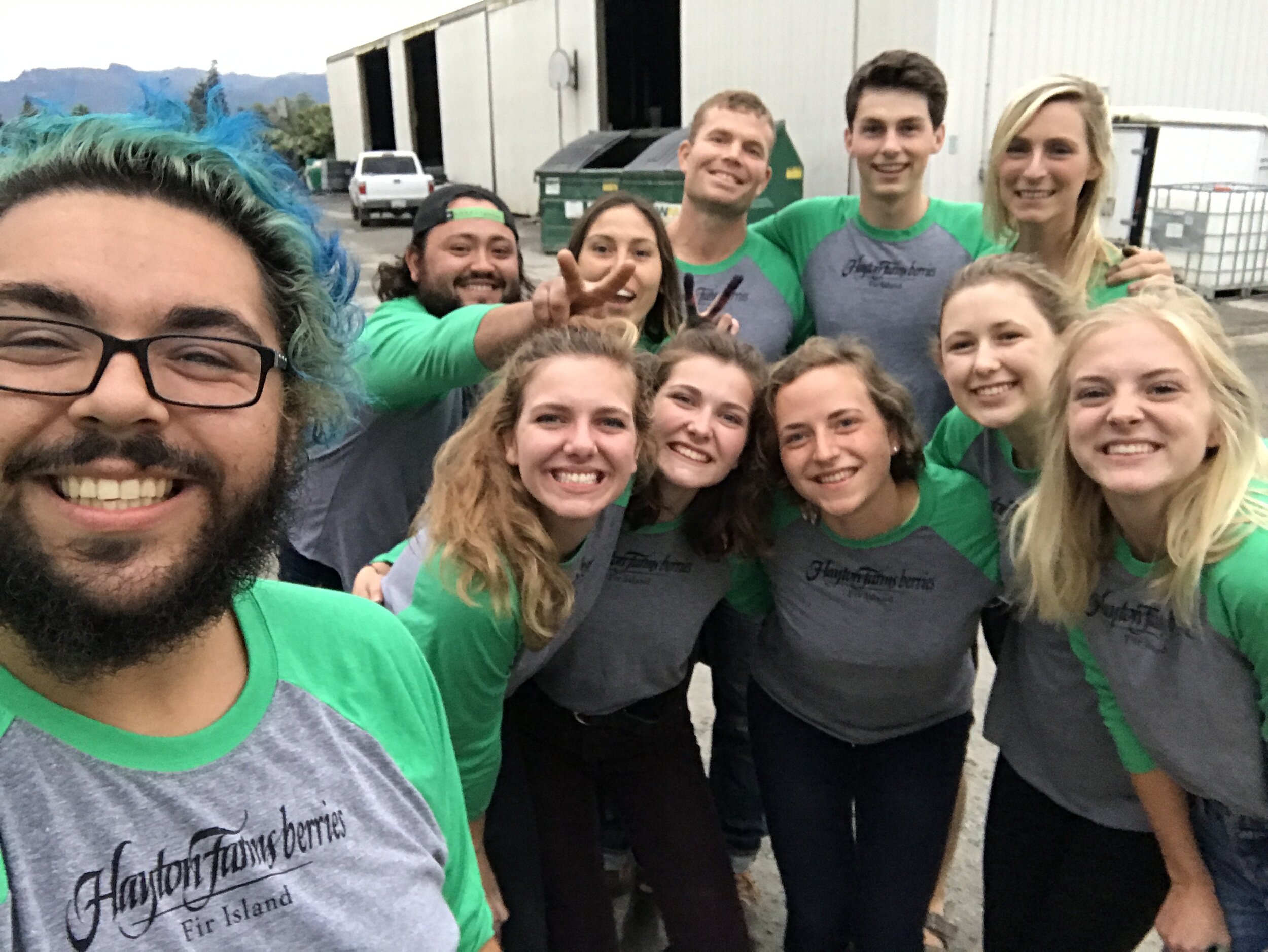 Group photo of cheerful team members wearing branded gray and green shirts from Hayton Farms Berries on Fir Island, standing outdoors near a warehouse with mountains in the background. The image captures a sense of camaraderie and teamwork.