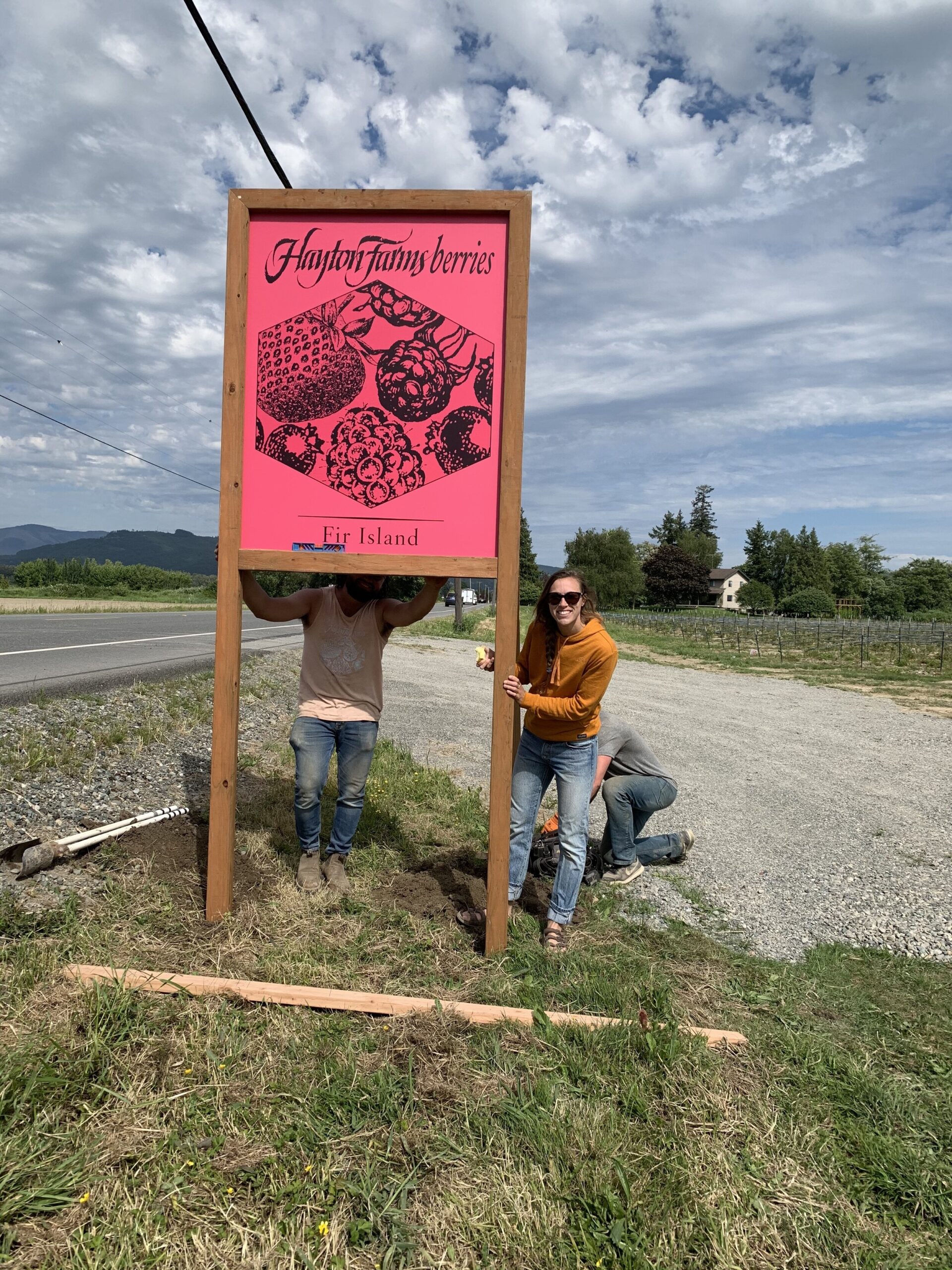 Two people holding a large pink sign for Hayton Farms Berries on Fir Island, with a scenic backdrop of fields and cloudy skies. The sign features illustrations of various berries and is part of the farm's branding.