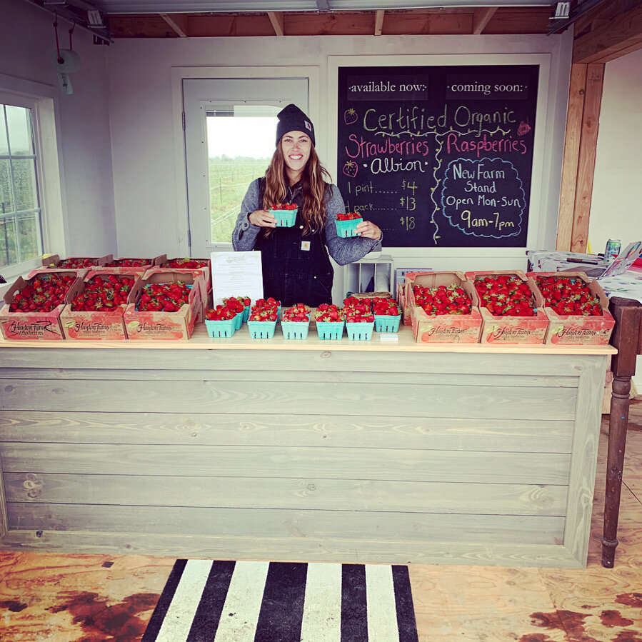 A young woman stands behind a rustic wooden table filled with fresh, certified organic strawberries in containers, showcasing the farm's produce. A chalkboard menu lists prices for strawberries and mentions upcoming availability of raspberries. The farm stand operates from 9 AM to 7 PM, open Monday through Sunday.