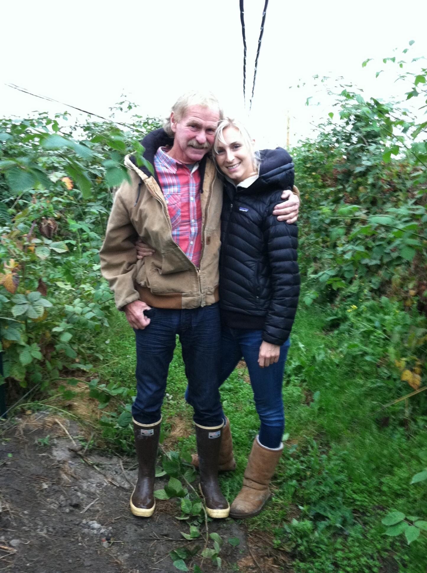 A smiling man and woman embrace in a lush garden setting, surrounded by green plants. The man wears a brown jacket, plaid shirt, and rubber boots, while the woman is dressed in a black puffer jacket and brown boots. The scene captures a warm, friendly moment in a natural environment.
