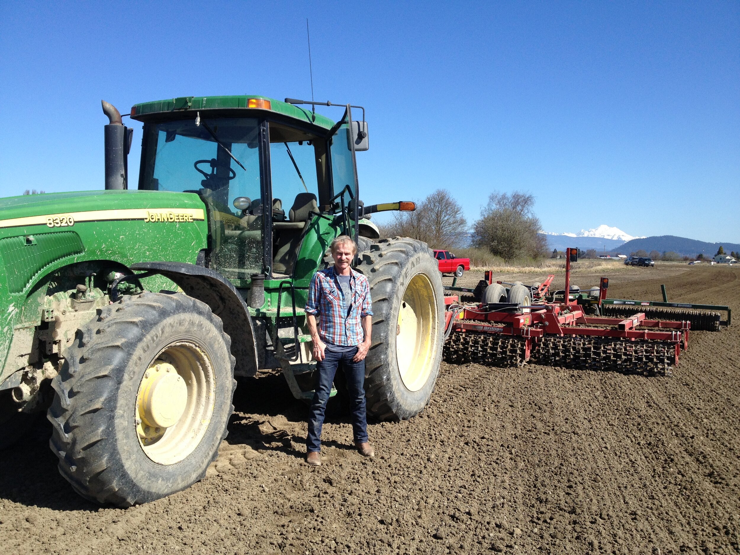 Farmer standing next to a John Deere tractor and tillage equipment on a plowed field with mountains in the background, showcasing agricultural practices in a sunny landscape.