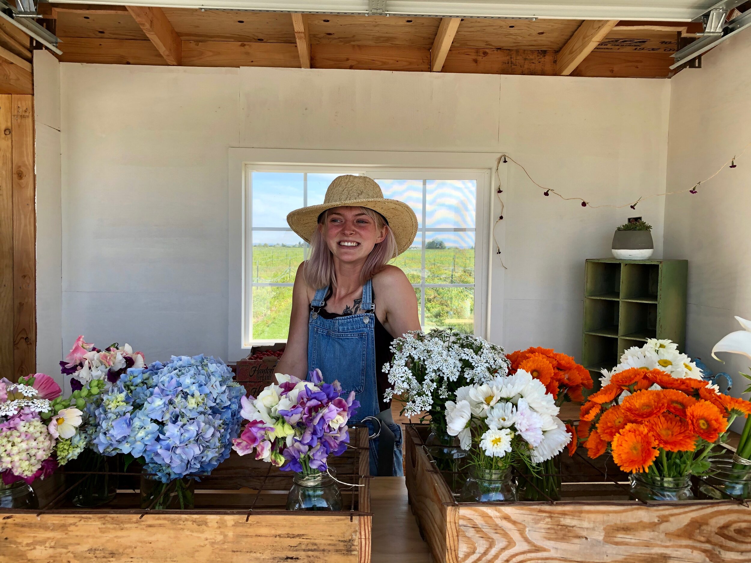 Smiling woman with pink hair and a straw hat stands behind a wooden flower stall filled with colorful bouquets including hydrangeas, daisies, and gerbera daisies, in a bright, airy space with a window view of fields.